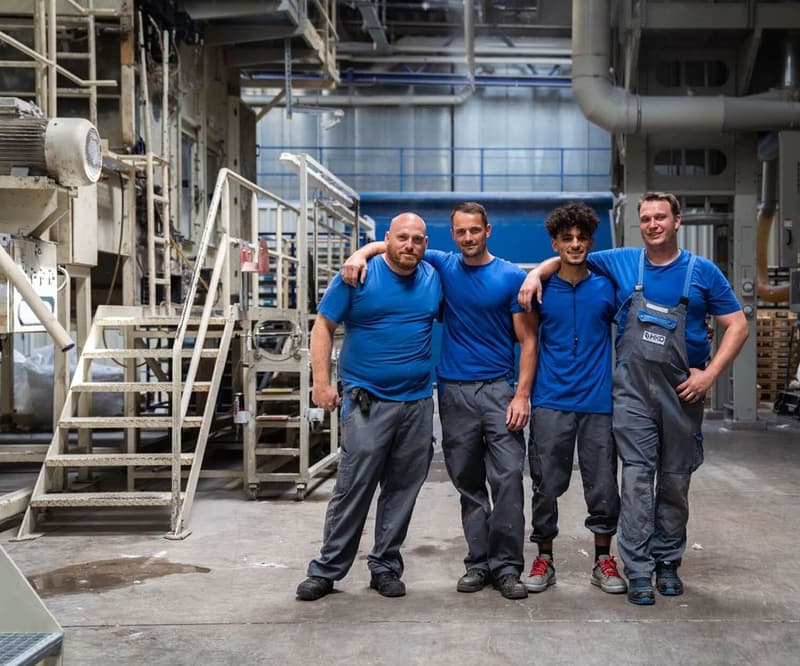 Vier Männer in blauen T-Shirts und Arbeitshosen stehen nebeneinander in einer Industriehalle