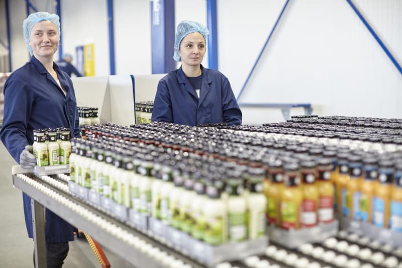 Zwei Mitarbeiterinnen mit Haarnetz und dunkelblauen Arbeitskitteln an einer Abfülllinie mit Flaschen